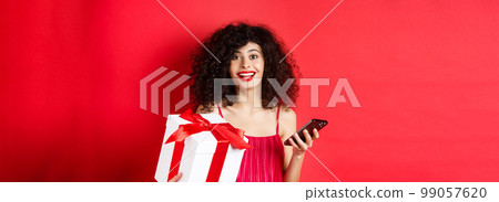 Online shopping and Valentines day. Beautiful young woman holding smartphone and lovers gift, looking surprised and happy at camera, red background 99057620