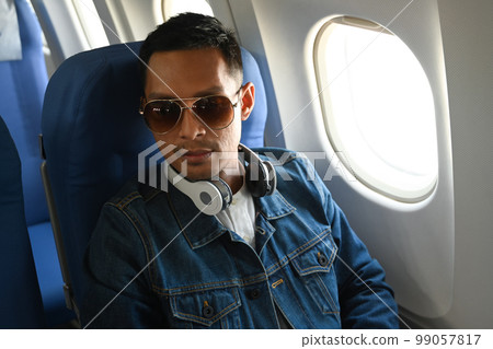 Hipster man passenger listening to music in wireless headphones while sitting in the airplane next to the window  Hipster man passenger listening to music in wireless headphones while sitting in the airplane next to the window  99057817