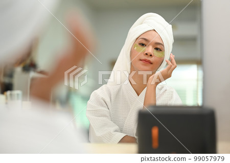 Charming millennial woman in bathrobe applying patches under eyes. Beauty treatment, skincare and cosmetics concept Charming millennial woman in bathrobe applying patches under eyes. Beauty treatment, skincare and cosmetics concept 99057979