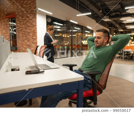 A red-haired caucasian woman uses a smartphone, and a bearded man rests at his desk in an open space office. 99058017
