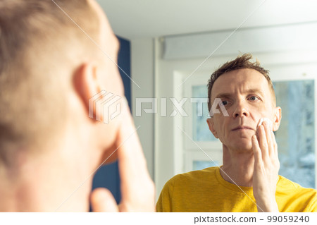 Mature man apply face tonic with cotton pad, then massage with hands. Male portrait in mirror. Face cleansing, washing. 99059240