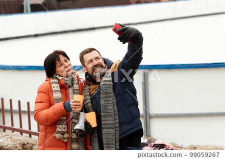Middle-aged man and woman, happy couple visiting open air ice rink in winter, taking photos, drinking hot tea. Positivity 99059267
