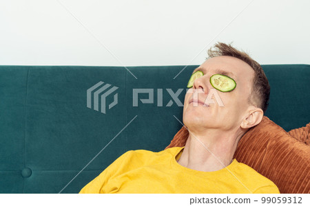 Happy man lying on sofa with eye patches from fresh cucumber slices in living room. Mask for dark circles under eyes. 99059312