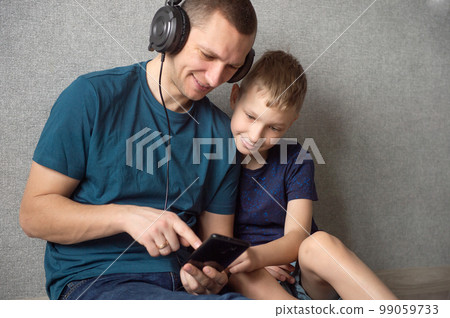 Dad is sitting on the floor next to his son with headphones on and pointing into the phone 99059733