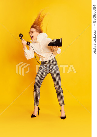 Excited girl with red hair wearing office style clothes shouting at retro vintage phone over yellow background. Concept of youth, student college life, business and education. 99060384