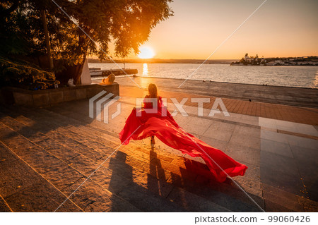 Sunrise red dress. A woman in a long red dress against the backdrop of sunrise, bright golden light of the sun's rays. The concept of femininity, harmony. 99060426