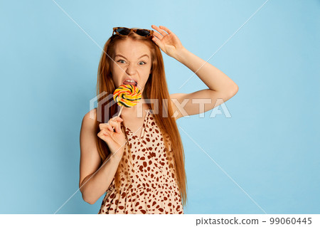 Portrait of emotional young beautiful redhaired girl with long glossy hair tasting lollipop and looking at camera isolated over light blue background. 99060445