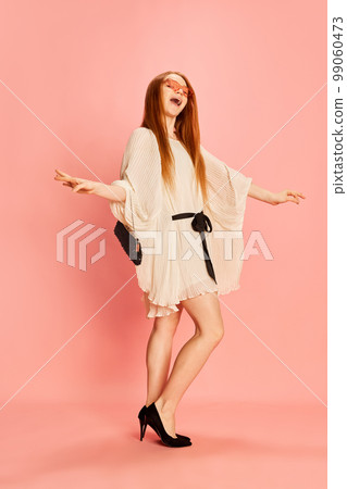 One young beautiful redhaired girl with long hair dancing, posing isolated over light pink background. Concept of positive emotions, happy mood, fashion, youth 99060473