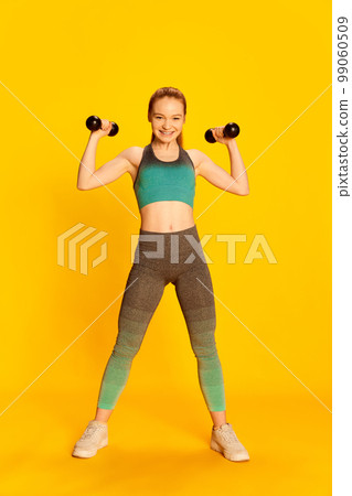 Strength. Happy beautiful girl in sports fitness clothes doing exercises with equipment over yellow background. Sport, fitness, happy positive emotions and active lifestyle concept. 99060509