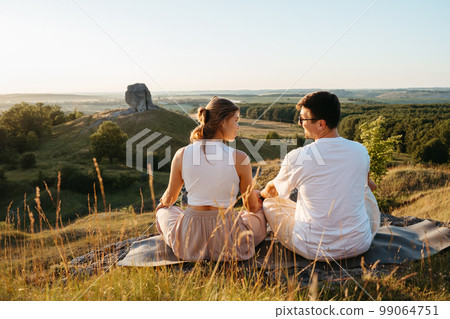 Man and Woman Practicing Yoga and Meditation Outdoors at Sunset with Scenic Landscape and Nature Miracle Giant Stone on the Background 99064751