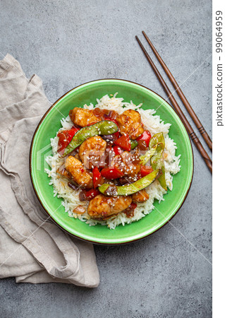 Asian sweet and sour sticky chicken with vegetables stir-fry and rice in ceramic bowl with chopsticks top view, gray rustic stone background, traditional Asian dish Asian sweet and sour sticky chicken with vegetables stir-fry and rice in ceramic bowl with chopsticks top view, gray rustic stone background, traditional Asian dish 99064959