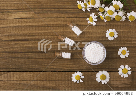 Homeopathic globules and glass bottle on a wooden natural background next to medicinal chamomile flowers. Homeopathic globules and glass bottle on a wooden natural background next to medicinal chamomile flowers. 99064998