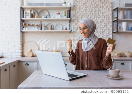 Happy female shopper in online store celebrating victory and successful purchase discounts, sales, muslim woman in hijab shopping online using laptop and bank credit card indoors at home in kitchen. Happy female shopper in online store celebrating victory and successful purchase discounts, sales, muslim woman in hijab shopping online using laptop and bank credit card indoors at home in kitchen. 99065131