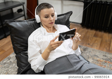 stylish caucasian adult woman at home wearing headphones watching video on smartphone stylish caucasian adult woman at home wearing headphones watching video on smartphone 99065914