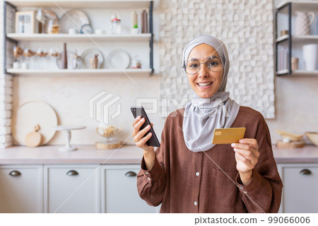 Portrait of happy online shopper woman at home, Muslim woman in hijab with phone smiling and looking at camera, Arab woman in kitchen holding bank credit card and phone. 99066006