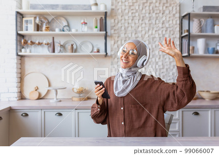 Happy and smiling woman dancing and singing at home in kitchen, Muslim woman in hijab and headphones listening to music and online radio smiling, using app on phone. 99066007