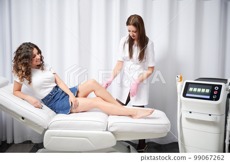 Doctor distributing gel on skin of client's shin with spatula for effective laser hair removal. Cosmetologist examining skin area of beautiful legs girl who undergoing laser hair removal. Doctor distributing gel on skin of client's shin with spatula for effective laser hair removal. Cosmetologist examining skin area of beautiful legs girl who undergoing laser hair removal. 99067262