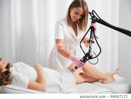 Dermatologist working with the latest laser device for polishing and repairing skin defects. Beautician demonstrating work of CO2 laser, warming skin of clients thigh, stimulating local rejuvenation. Dermatologist working with the latest laser device for polishing and repairing skin defects. Beautician demonstrating work of CO2 laser, warming skin of clients thigh, stimulating local rejuvenation. 99067272
