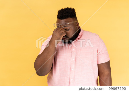 Portrait of frustrated man wearing pink t shirt and optical glasses touching closed eyes, grief or pain after long hours working, feeling hopeless. Indoor studio shot isolated on yellow background. 99067290
