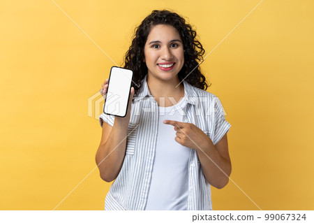 Portrait of smiling attractive young adult woman with dark wavy hair holding pointing at cell phone with empty display, copy space for advertisement. Indoor studio shot isolated on yellow background. Portrait of smiling attractive young adult woman with dark wavy hair holding pointing at cell phone with empty display, copy space for advertisement. Indoor studio shot isolated on yellow background. 99067324