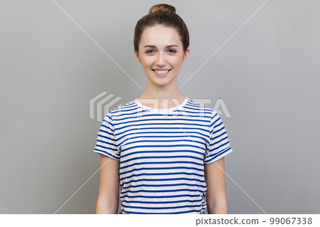 Portrait of attractive charming woman wearing striped T-shirt standing looking at camera with toothy smile, expressing happiness and positive emotions. Indoor studio shot isolated on gray background. 99067338