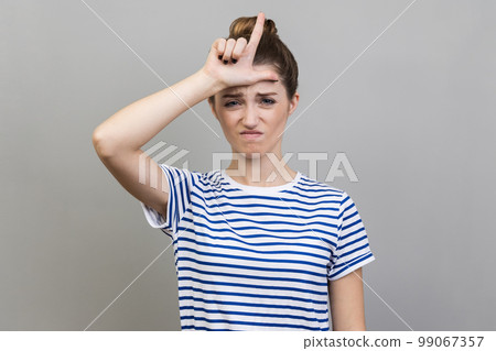 Portrait of upset disappointed woman wearing striped T-shirt standing, showing looser gesture with hand on forehead, looking at camera. Indoor studio shot isolated on gray background. 99067357