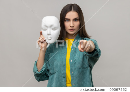 Portrait of bossy woman with dark hair holding white mask in hand, pointing finger to camera with serious expression, wearing casual style jacket. Indoor studio shot isolated on gray background. 99067362