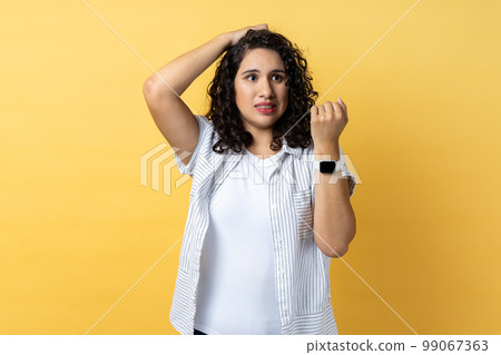 Portrait of beautiful adorable confused woman with dark wavy hair standing with puzzled facial expression, looks pensive, showing smart watch. Indoor studio shot isolated on yellow background. Portrait of beautiful adorable confused woman with dark wavy hair standing with puzzled facial expression, looks pensive, showing smart watch. Indoor studio shot isolated on yellow background. 99067363