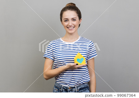 Portrait of pretty woman wearing striped T-shirt holding in hands little paper house looking at camera, dreaming about her own apartment. Indoor studio shot isolated on gray background. Portrait of pretty woman wearing striped T-shirt holding in hands little paper house looking at camera, dreaming about her own apartment. Indoor studio shot isolated on gray background. 99067364