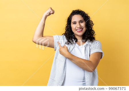 Portrait of woman with dark wavy hair showing her arm muscle and point it with finger, proud of her strength and leadership skills. Indoor studio shot isolated on yellow background. 99067371
