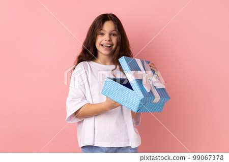 Portrait of little girl wearing white T-shirt opening gift box and looking at camera with excited facial expression, satisfied with present. Indoor studio shot isolated on pink background. Portrait of little girl wearing white T-shirt opening gift box and looking at camera with excited facial expression, satisfied with present. Indoor studio shot isolated on pink background. 99067378