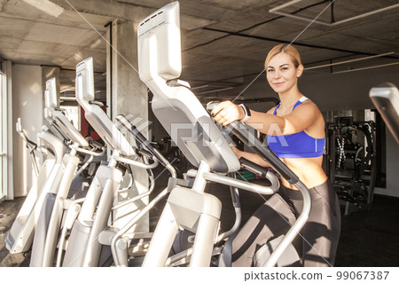 Portrait of young fit athletic woman using elliptic trainer in a fitness center, looking at camera and smiling, wearing sports top and tights. Indoor shot with window on background. 99067387