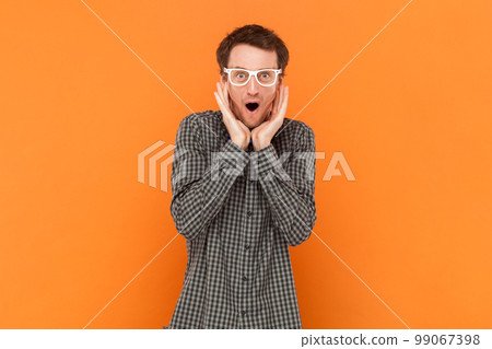 Amazed man nerd doesnt believe his success, keeps hands on head, stares at camera, says omg or wow, wearing shirt with blue bow tie and white glasses. Indoor studio shot isolated on orange background Amazed man nerd doesnt believe his success, keeps hands on head, stares at camera, says omg or wow, wearing shirt with blue bow tie and white glasses. Indoor studio shot isolated on orange background 99067398