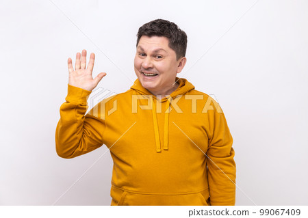 Friendly positive man greeting you rising hand and waving, saying hi, glad to see you, looking at camera with toothy smile, wearing urban style hoodie. Indoor studio shot isolated on white background. 99067409
