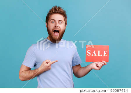 Portrait of excited amazed bearded man pointing finger at card with sale word, opening mouth in amazement, looking shocked by discounts. Indoor studio shot isolated on blue background. Portrait of excited amazed bearded man pointing finger at card with sale word, opening mouth in amazement, looking shocked by discounts. Indoor studio shot isolated on blue background. 99067414