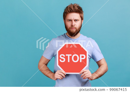 Portrait of handsome serious young adult bearded man holding octagonal Stop symbol, showing red traffic sign, warning of restricted area. Indoor studio shot isolated on blue background. Portrait of handsome serious young adult bearded man holding octagonal Stop symbol, showing red traffic sign, warning of restricted area. Indoor studio shot isolated on blue background. 99067415