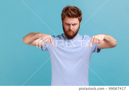 Here and right now. Portrait of bearded man standing and looking at camera with fingers down, commanding to act here and now, demanding immediate result. Indoor studio shot isolated on blue background Here and right now. Portrait of bearded man standing and looking at camera with fingers down, commanding to act here and now, demanding immediate result. Indoor studio shot isolated on blue background 99067417