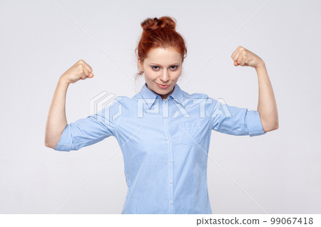 Portrait of strong confident young adult ginger woman wearing blue shirt raising arms showing biceps, female strength and feminism. Indoor studio shot isolated on gray background. Portrait of strong confident young adult ginger woman wearing blue shirt raising arms showing biceps, female strength and feminism. Indoor studio shot isolated on gray background. 99067418
