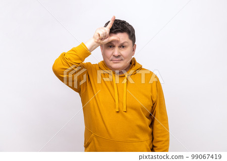 Gloomy depressed man showing looser gesture holding fingers near forehead, sad because of silly mistake, wearing urban style hoodie. Indoor studio shot isolated on white background. 99067419
