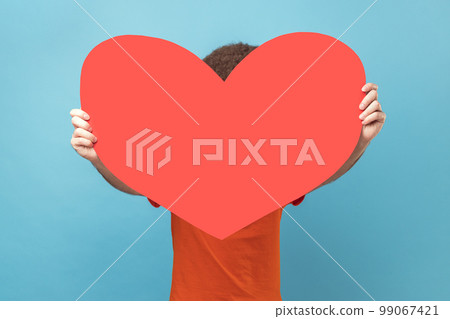 Portrait of man wearing orange T-shirt hiding his face behind big red paper heart, making anonymous surprise, sending greeting card. Indoor studio shot isolated on blue background. Portrait of man wearing orange T-shirt hiding his face behind big red paper heart, making anonymous surprise, sending greeting card. Indoor studio shot isolated on blue background. 99067421