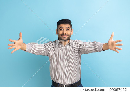 Come into my arms. Portrait of good natured businessman with beard reaching out to camera, stretching arms to hug you, wearing striped shirt. Indoor studio shot isolated on blue background. Come into my arms. Portrait of good natured businessman with beard reaching out to camera, stretching arms to hug you, wearing striped shirt. Indoor studio shot isolated on blue background. 99067422