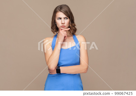 Portrait of attractive thoughtful puzzled woman with wavy hair holding chin, looking away with pensive expression, planning, wearing blue dress. Indoor studio shot isolated on light brown background. 99067425