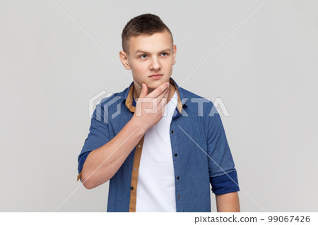 Portrait of serious pensive handsome teenager guy wearing blue shirt looking away, holding chin, thinking about future plans, planning. Indoor studio shot isolated on gray background. 99067426