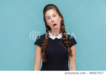 Portrait of naughty woman with braids sticking out tongue, making funny grimace, looking and winking at camera, wearing black dress. woman Indoor studio shot isolated on blue background. Portrait of naughty woman with braids sticking out tongue, making funny grimace, looking and winking at camera, wearing black dress. woman Indoor studio shot isolated on blue background. 99067427