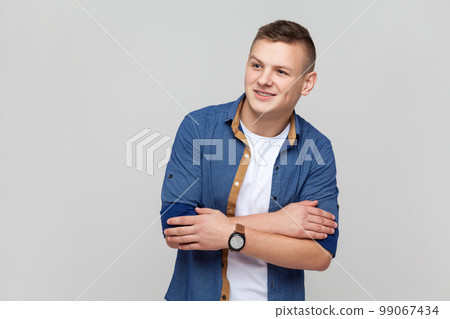 Portrait of handsome attractive young smiling teenager boy wearing blue shirt keeps hands folded, looking away with dreaming expression. Indoor studio shot isolated on gray background. Portrait of handsome attractive young smiling teenager boy wearing blue shirt keeps hands folded, looking away with dreaming expression. Indoor studio shot isolated on gray background. 99067434