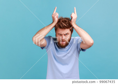 Portrait of angry bully bearded man showing bull horn gesture with fingers over head, looking hostile and threatening, aggressive face. Indoor studio shot isolated on blue background. 99067435