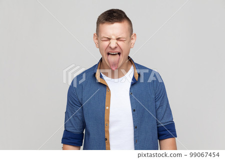 Childish disobedient behavior. Portrait of attractive teenager guy wearing blue shirt sticking tongue out, making naughty goofy grimace, teasing. Indoor studio shot isolated on gray background. 99067454