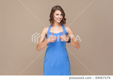 Portrait of attractive positive optimistic woman with wavy hair standing with raised thumbs up, recommended service, wearing blue dress. Indoor studio shot isolated on light brown background. 99067479