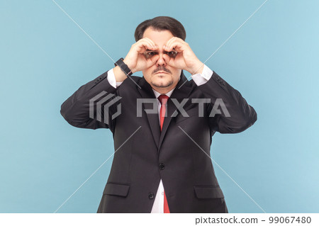 Portrait of serious concentrated man with mustache looking at camera through binocular making with hands, wearing black suit with red tie. Indoor studio shot isolated on light blue background. 99067480