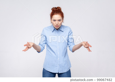 Portrait of unhappy frustrated ginger woman wearing blue shirt spreading hands with shock and misunderstanding, how could you sign. Indoor studio shot isolated on gray background. 99067482
